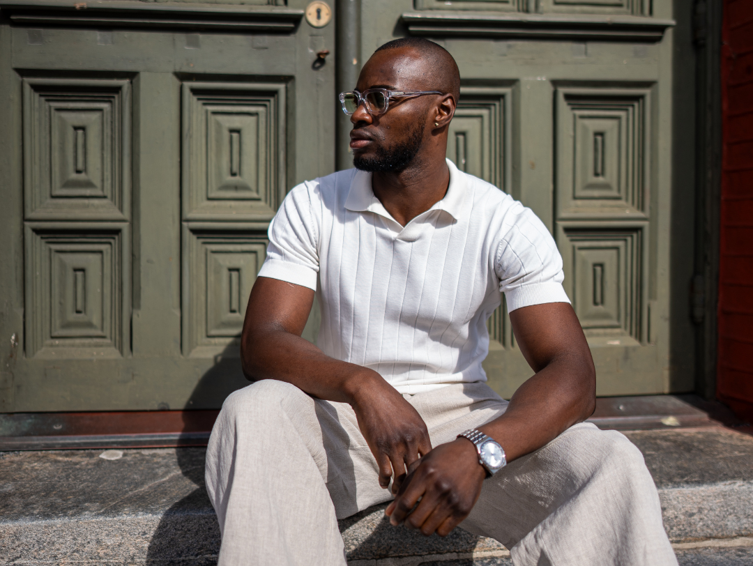 Man wearing sunglasses in a white polo shirt, white tone outfit sitting next to a green door.