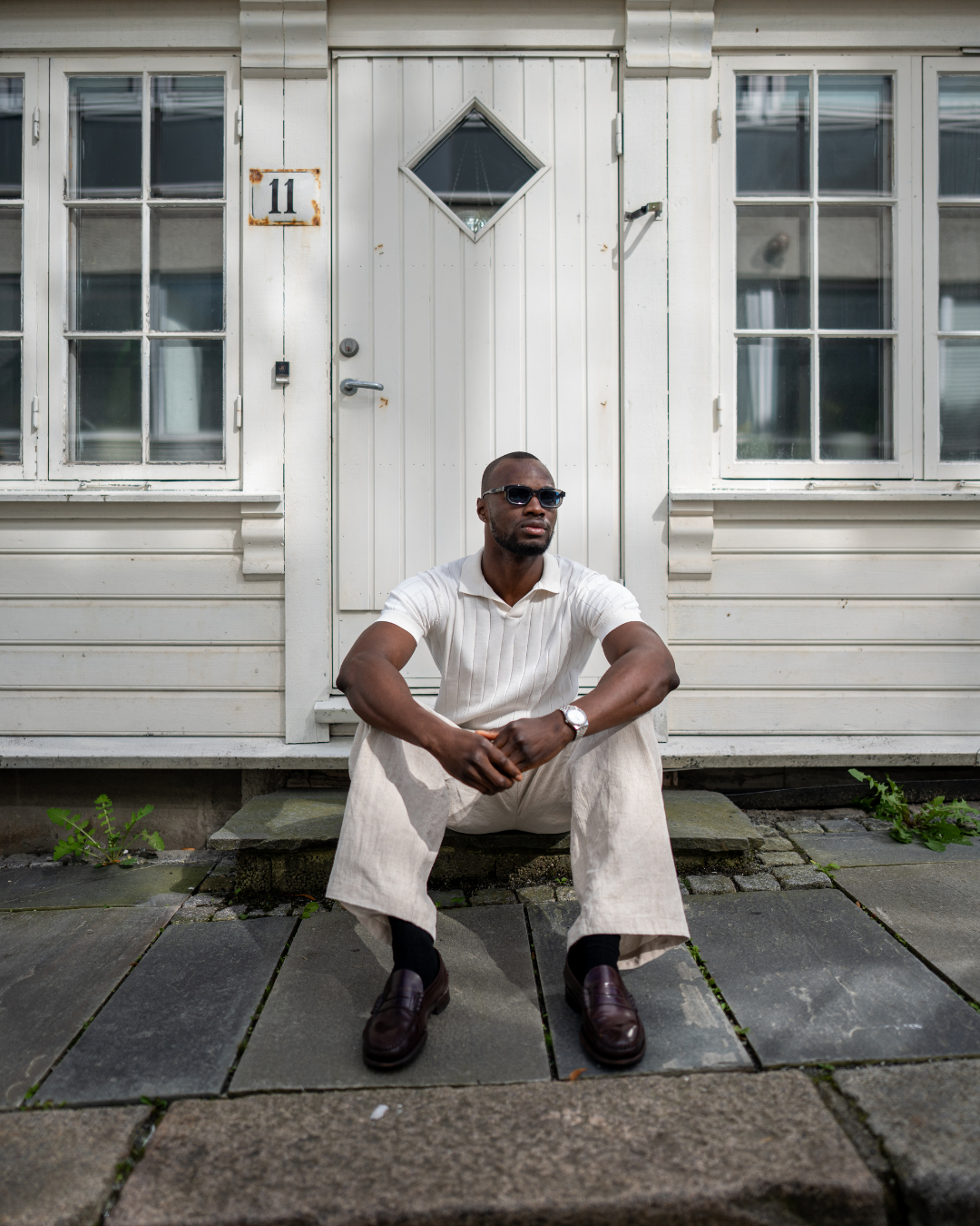 Man sitting on a stone step in front of a white wooden house, wearing the UR3328 Bleu sunglasses