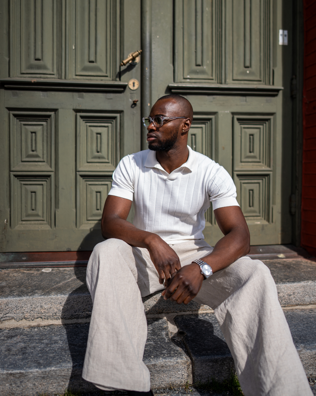 Man wearing sunglasses in a white polo shirt, white tone outfit sitting next to a green door.