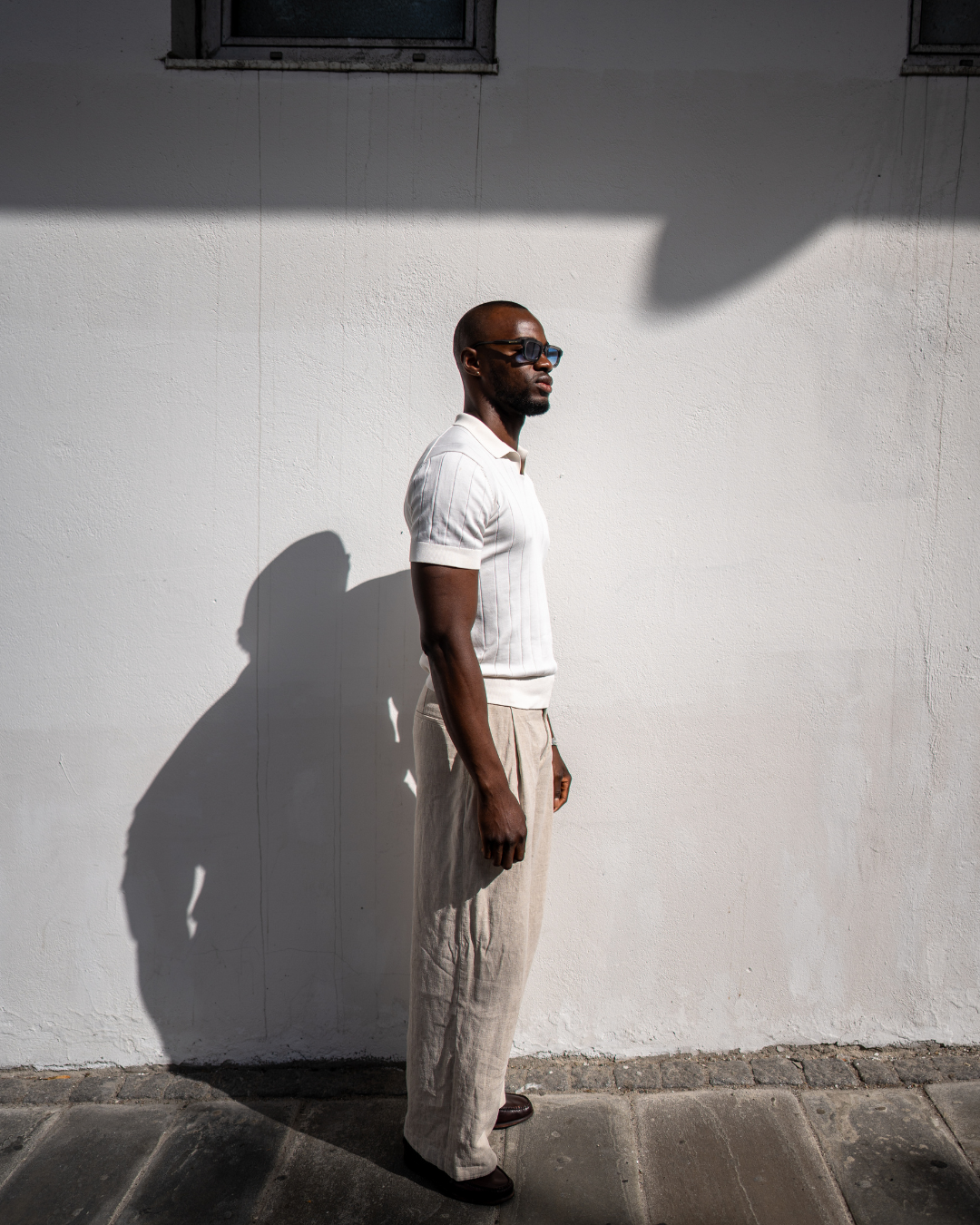 Man wearing sunglasses with a white polo shirt, white tone linnen pants and brown loafers standing next to a white wall.